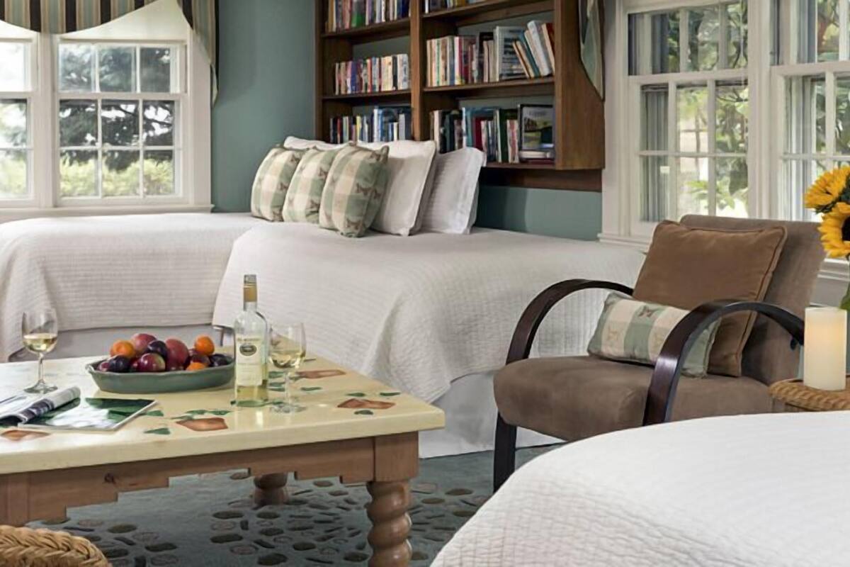 A cozy bedroom with two beds, a bedside table with a bowl of fruit, a bottle of wine, and a lamp.