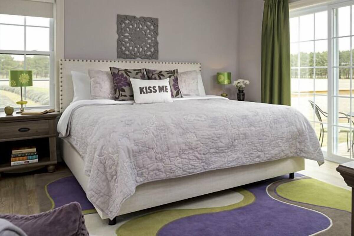 A bedroom with a large bed, a nightstand, a bedside table, a window with curtains, and a view of the outdoors.