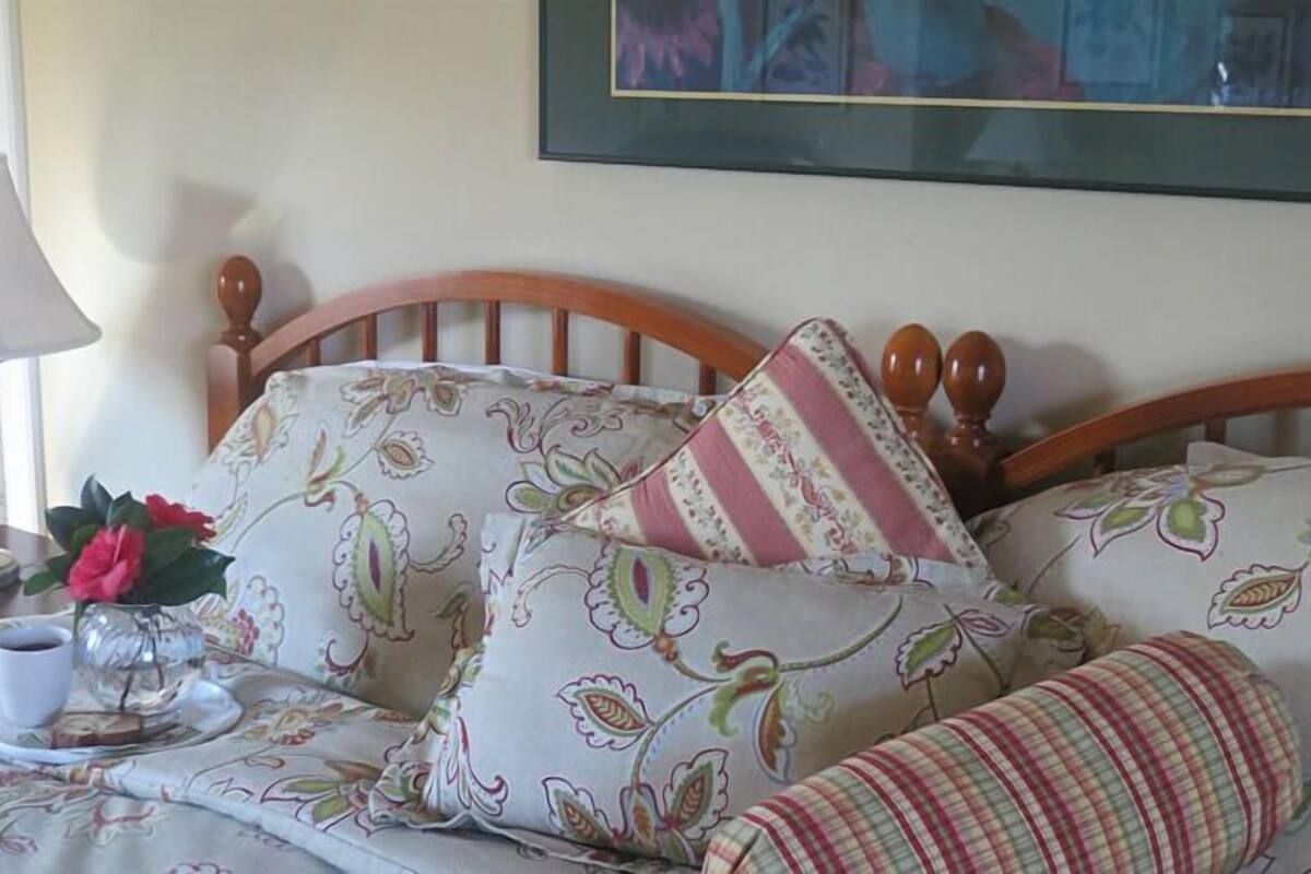 A neatly made bed with floral patterned bedding, a bedside table with a lamp and a cup of coffee, and a framed artwork on the wall.