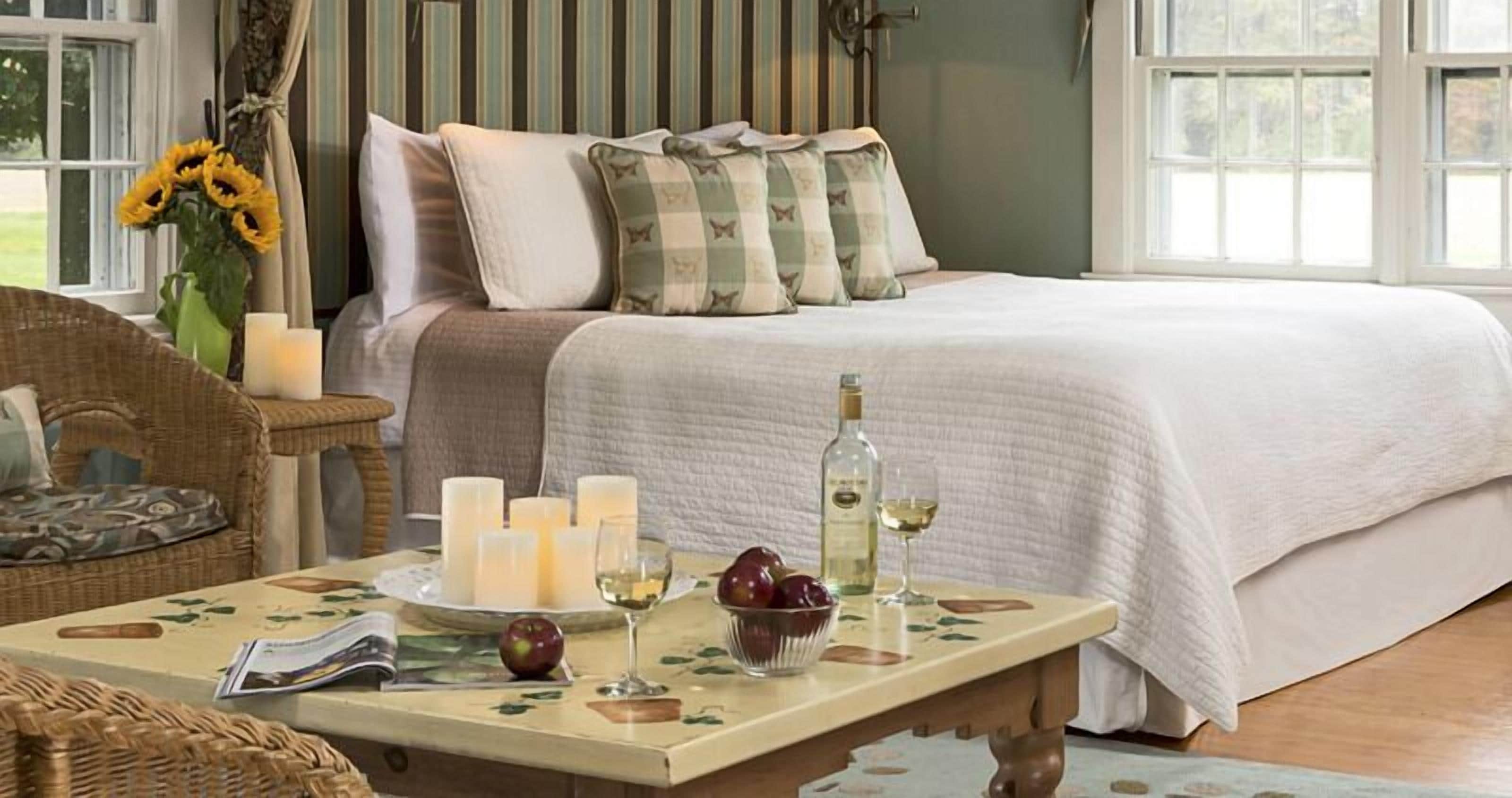 A bedroom with a bed, bedside table with candles, a vase with sunflowers, and a table with apples and wine glasses.