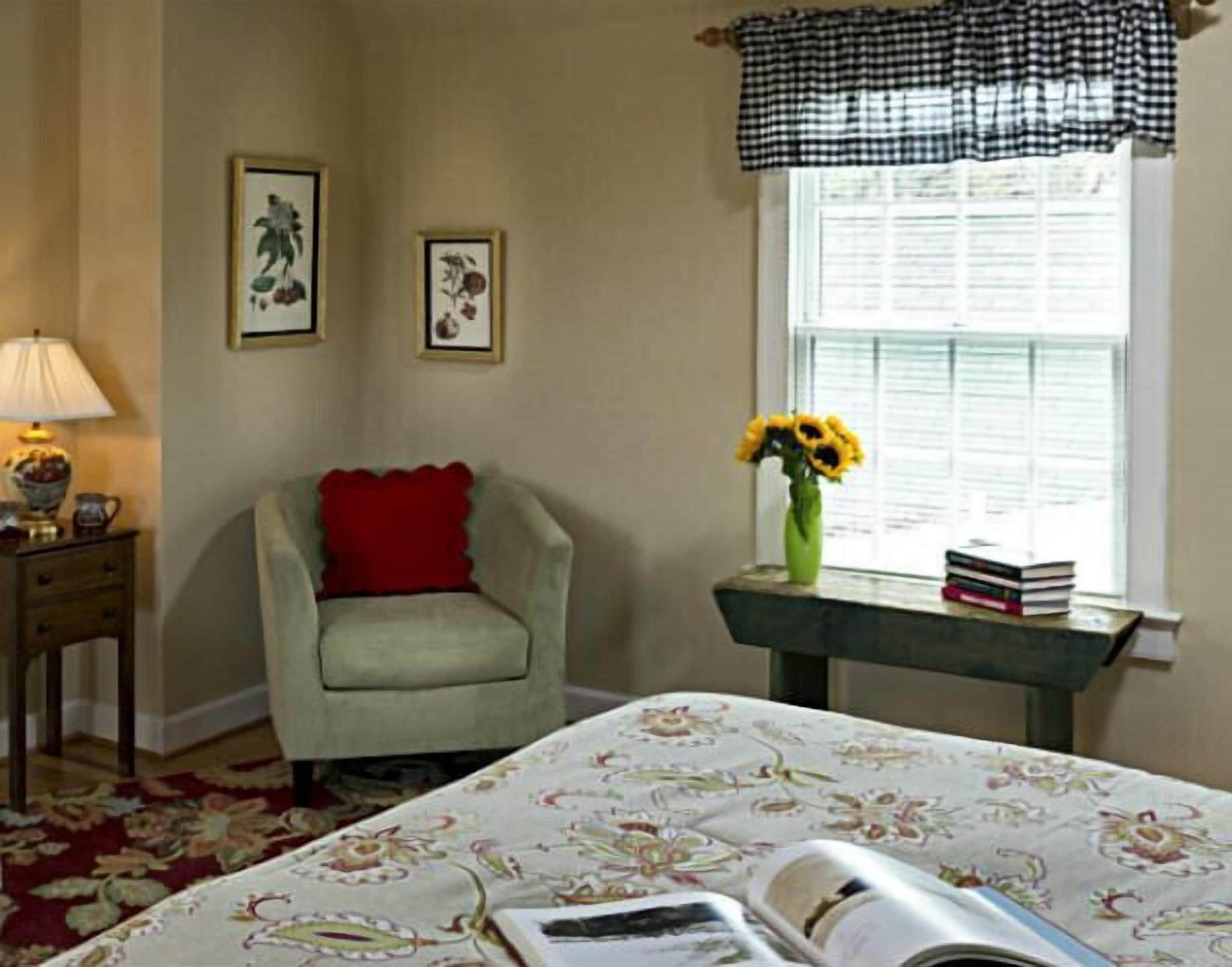 Een kamer met een bed, een stoel, een tafel met boeken en een bloevas, en twee ingelijste foto's aan de muur.