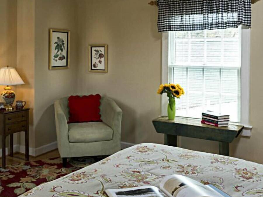 Een kamer met een bed, een stoel, een tafel met boeken en een bloevas, en twee ingelijste foto's aan de muur.