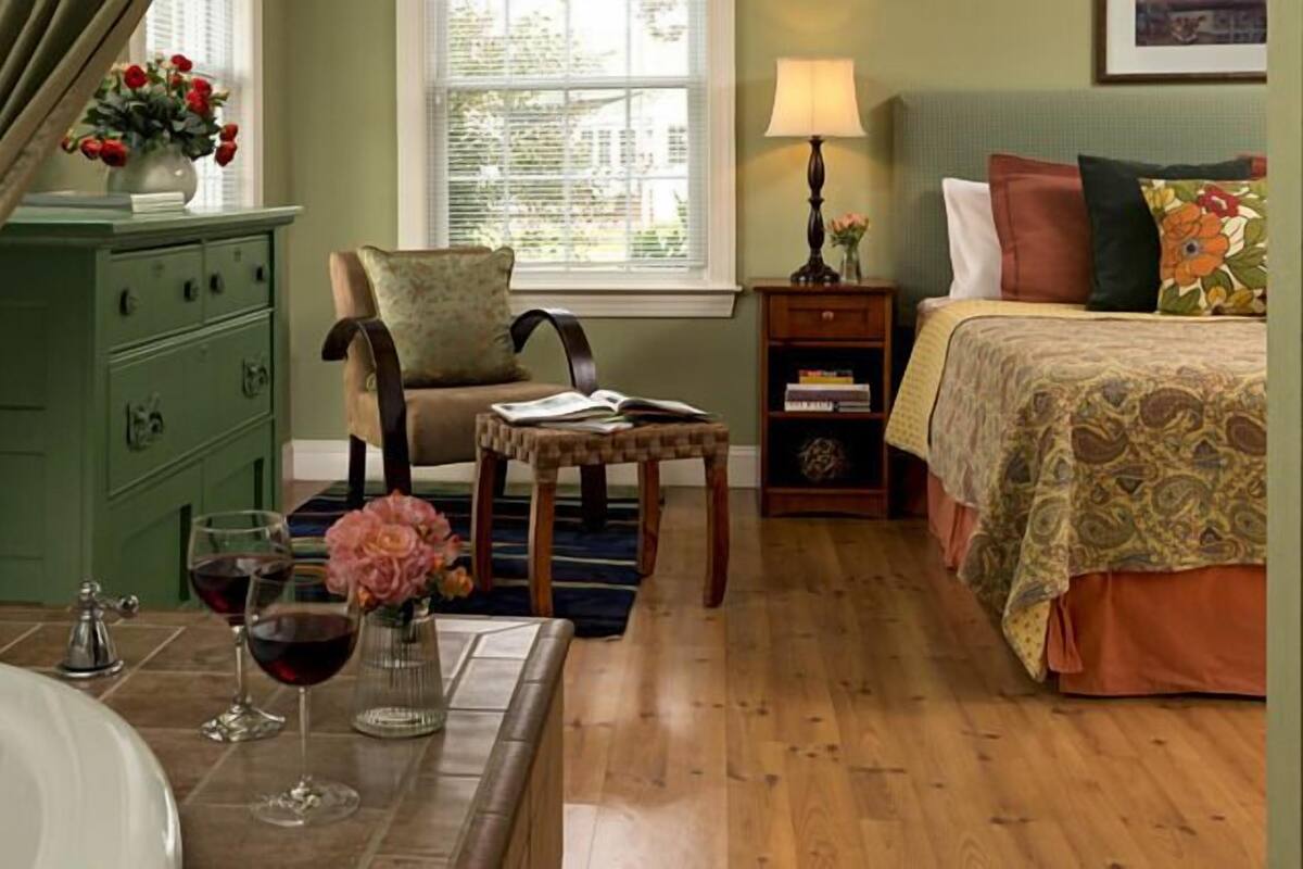 A room with a bed, a chair, a small table, a dresser, a lamp, and a window with blinds.
