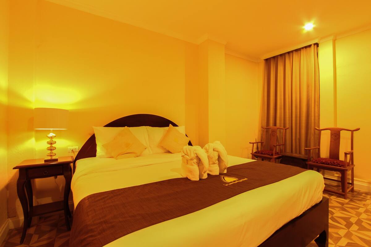 A hotel room with a large bed, a nightstand with a lamp, a wooden chair, and a window with curtains.