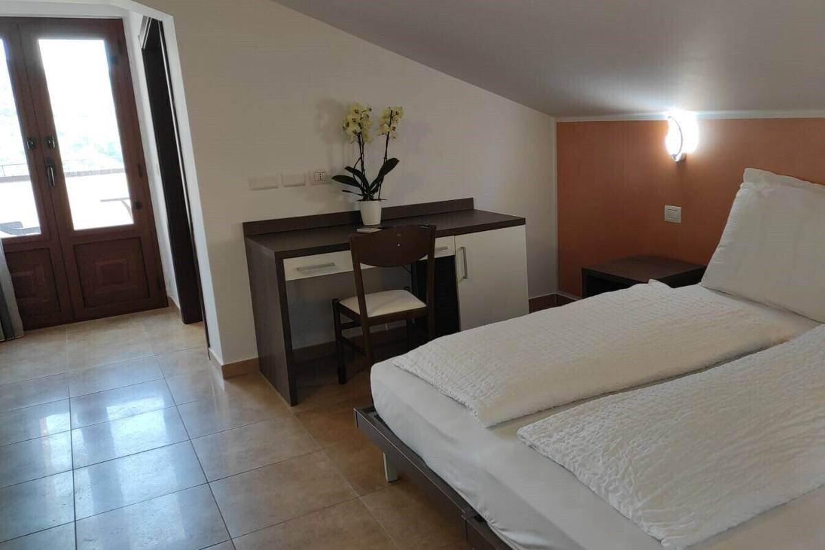 Ein Hotelzimmer mit einem Bett, einem Schreibtisch mit Stuhl, einem Nachttisch, einer Wandleuchte und einer Zimmerpflanze.