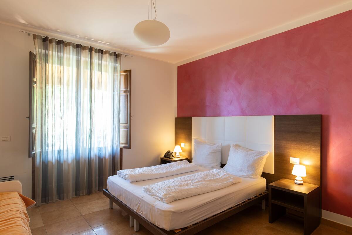 Ein Hotelzimmer mit einem Bett, zwei Nachttischen mit Lampen, einem Fenster mit transparenten Vorhängen und einer roten Akzentwand.