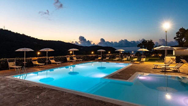 A swimming pool with lounge chairs and umbrellas, surrounded by a landscaped area with a view of a hilly landscape. - Villa Ginevra Resort Agrituristico (Ficarra)