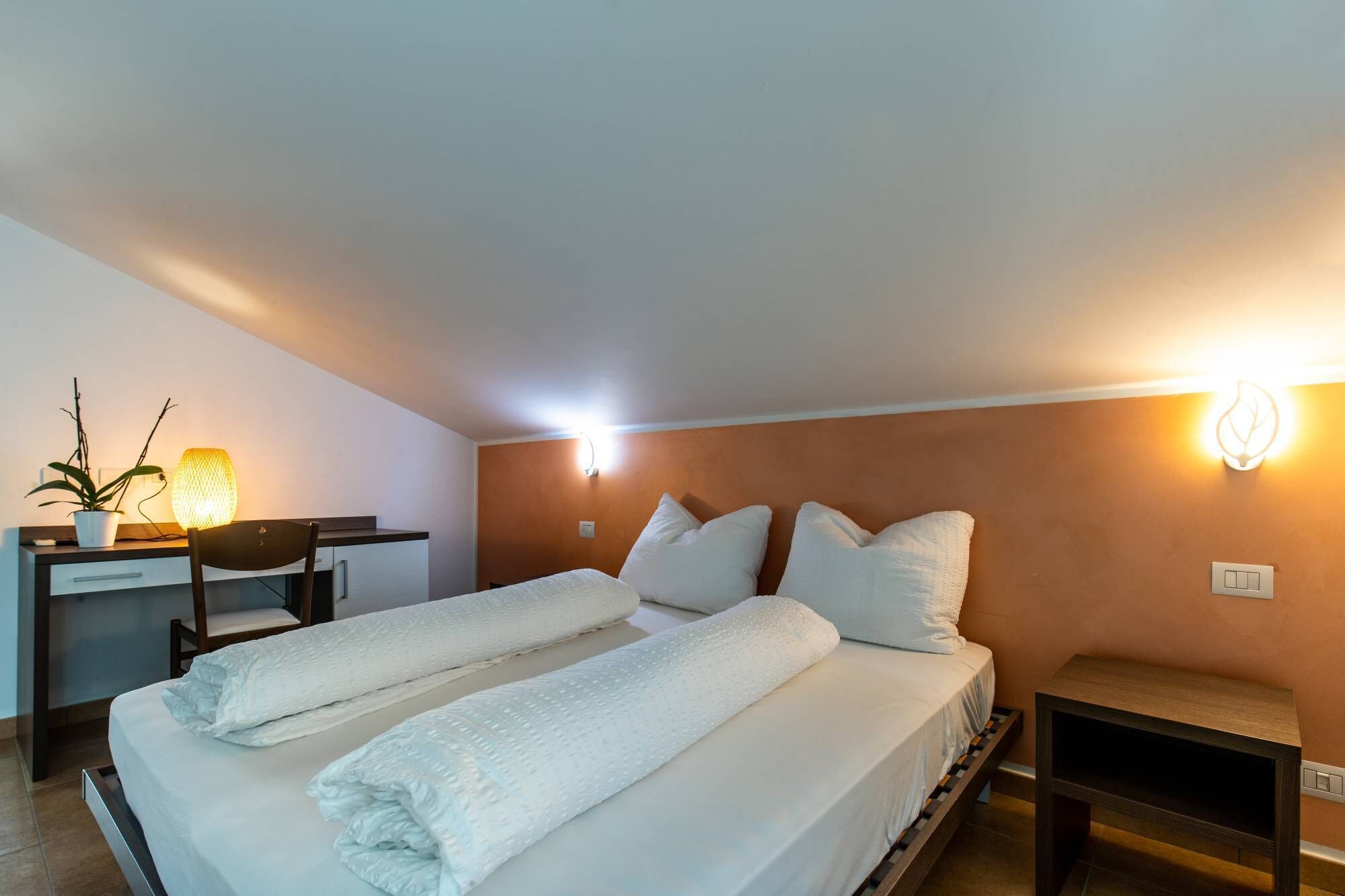 Ett hotellrum med en säng, sängbord, ett skrivbord, en stol och en väggmonterad lampa.