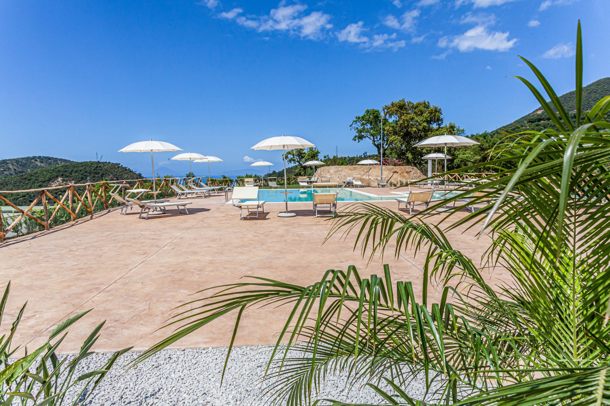 A pool area with lounge chairs and umbrellas, surrounded by greenery and hills.