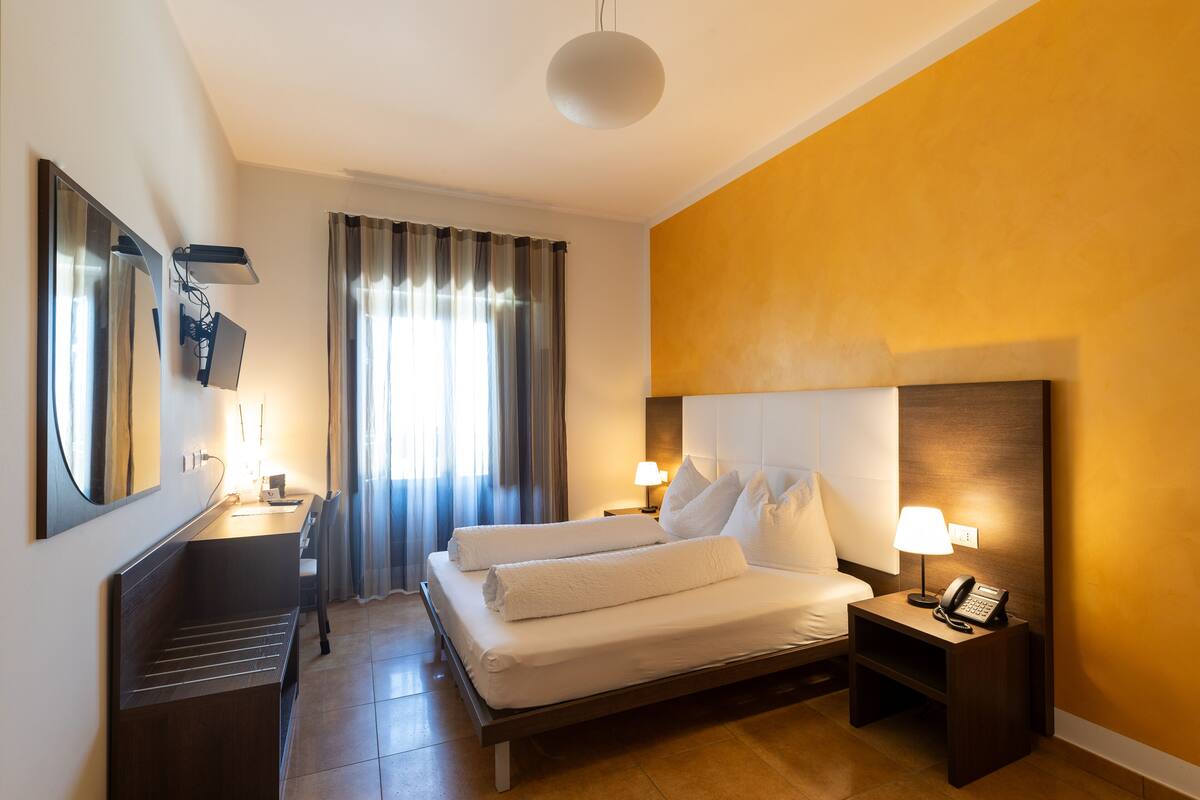 Ein Hotelzimmer mit Bett, Nachttisch mit Lampe, Schreibtisch mit Stuhl, Spiegel und einem Fenster mit Vorhängen.