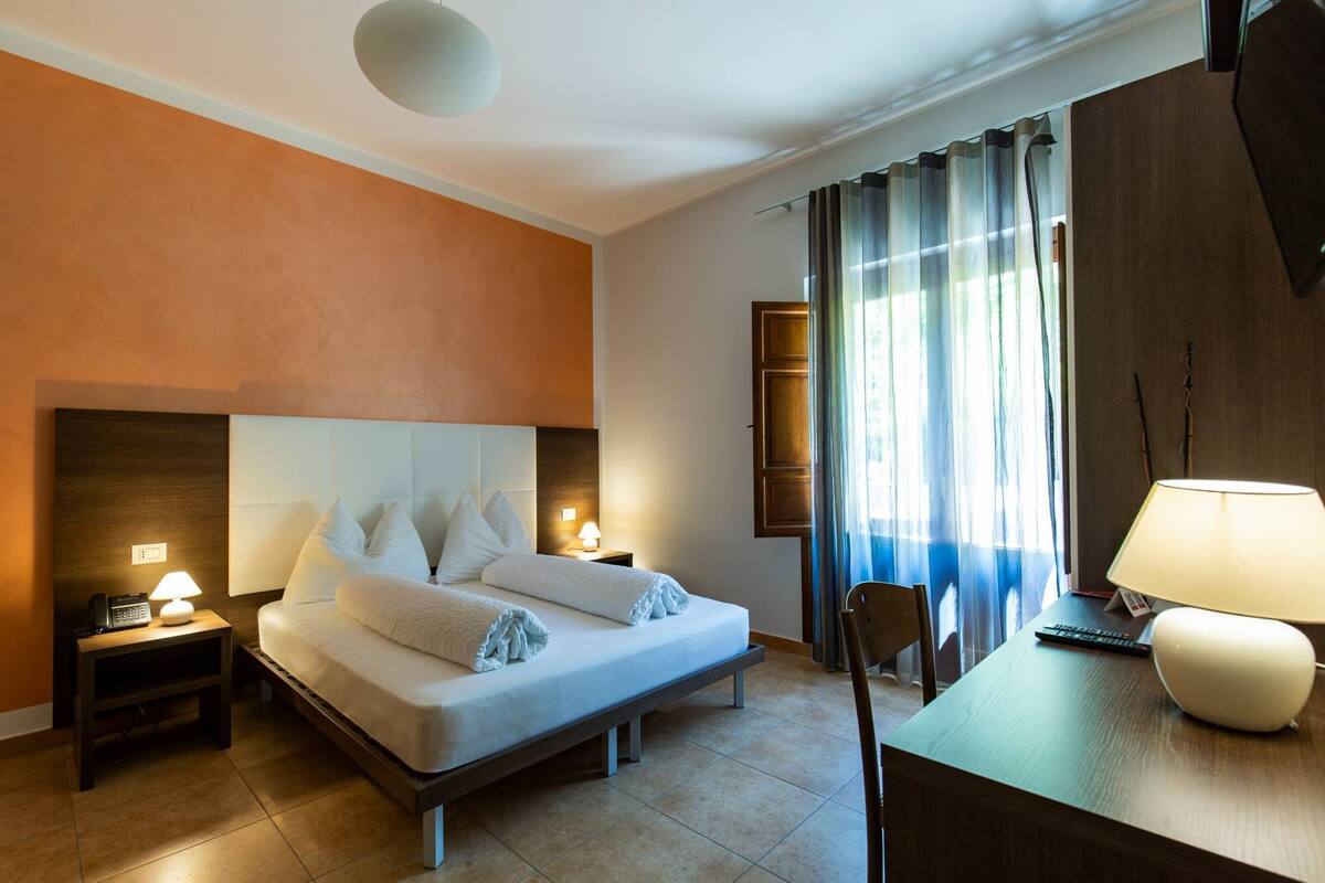 Ein Hotelzimmer mit Bett, Nachttischen mit Lampen, Schreibtisch mit Lampe, Stuhl und einem fenster mit Vorhängen.