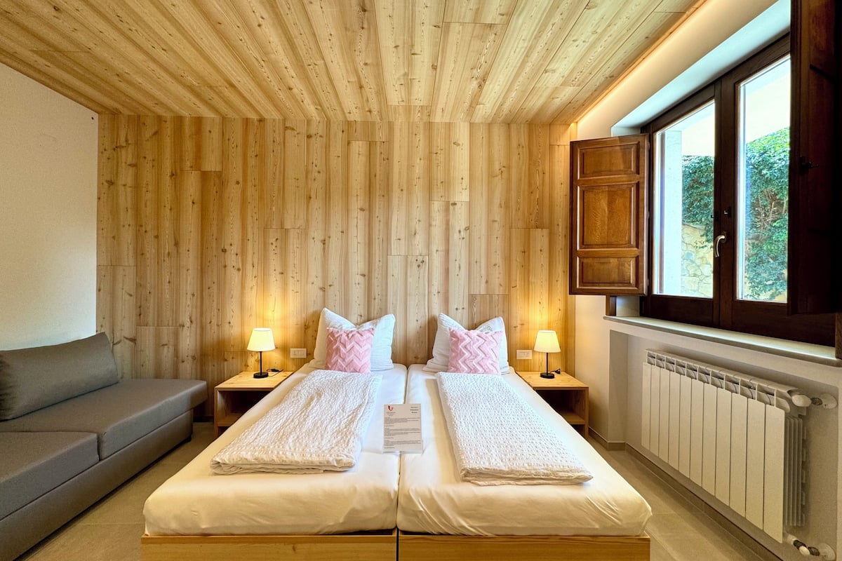 Ein Schlafzimmer mit Holzboden, zwei Betten mit weißer Bettwäsche, Nachttischen mit Lampen, einer Holzbank und einem Fenster mit Blick auf Grünflächen.