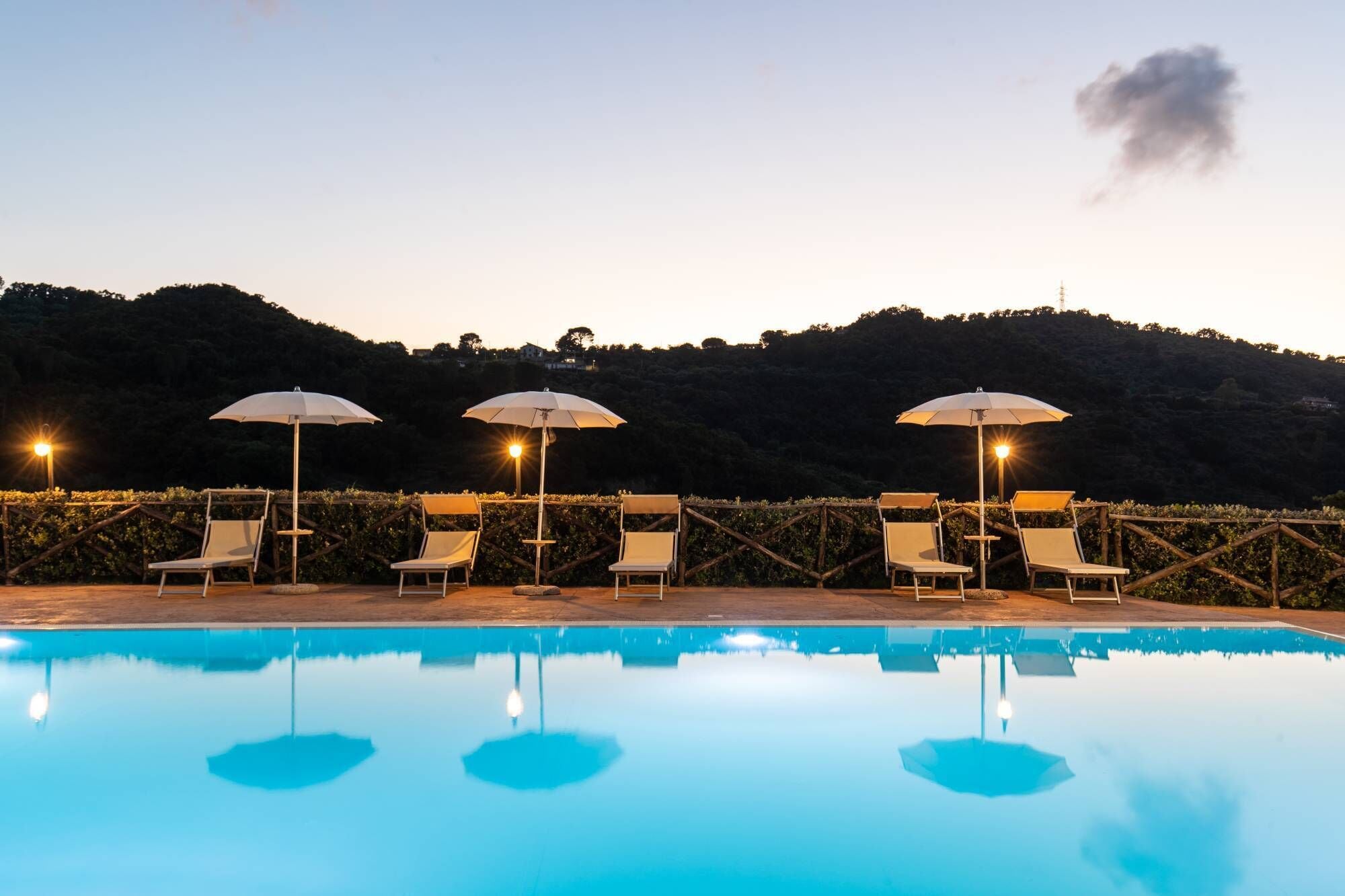 A poolside area with lounge chairs and umbrellas, surrounded by a wooden fence and a hillside in the background.