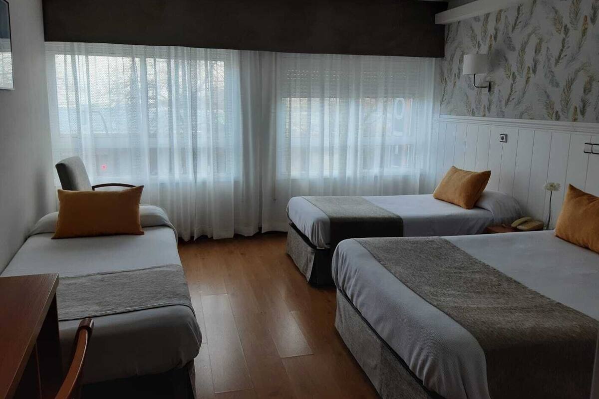 Una habitación de hotel con dos camas, una silla, un escritorio y una ventana con cortinas translúcidas.
