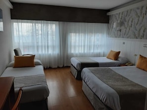 Ein Hotelzimmer mit zwei Betten, einem Sessel, einem Schreibtisch und einem Fenster mit durchsichtigen Vorhängen.