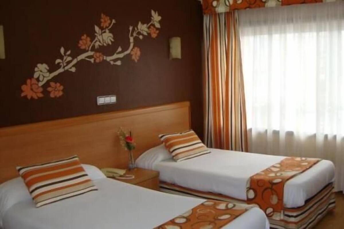 Habitación de hotel con dos camas, cabecera de madera, mesita de noche, una silla y una ventana con cortinas.