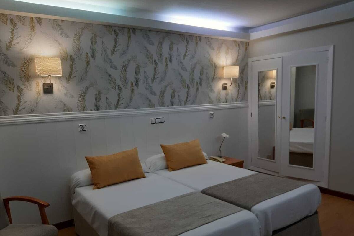 Una cama doble con dos almohadas, una mesita de noche, una silla y un espejo en una habitación de hotel.