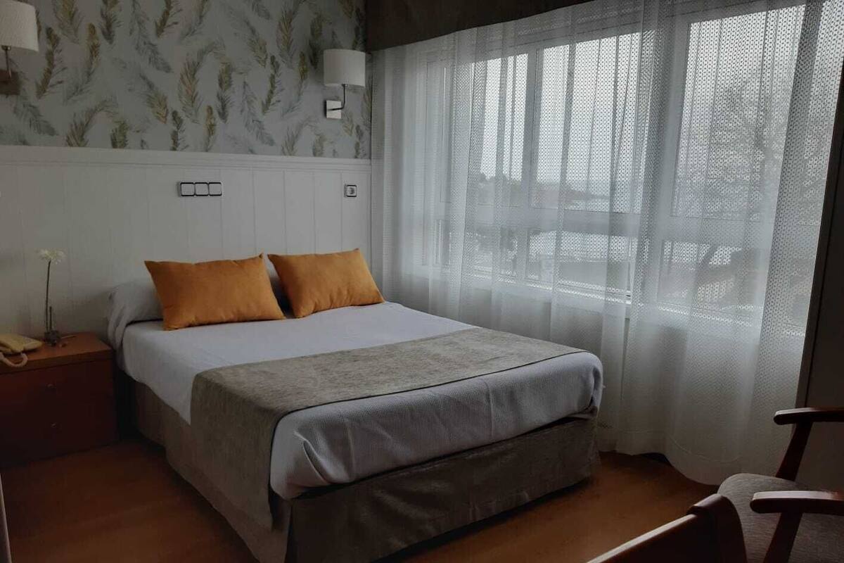 Un dormitorio ordenado con cama, mesita de noche, una silla y ventana con cortinas de gasa.