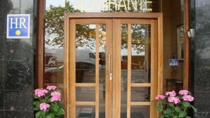 Entrada de hotel con puerta giratoria, un cartel que muestra 'HR' y un símbolo de sol, y flores rosadas al frente.