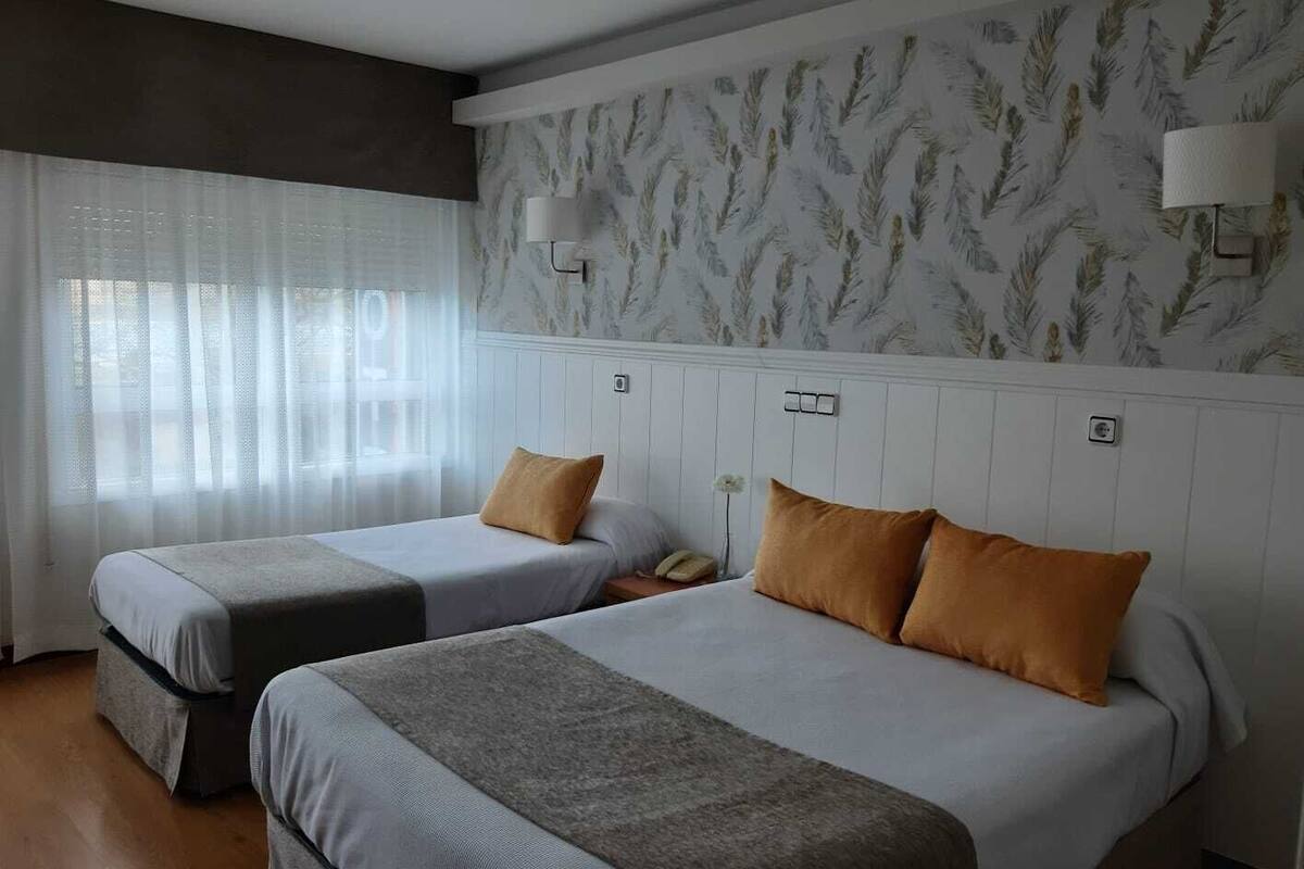 Habitación de hotel con dos camas, un ventanal con cortinas translúcidas y un papel tapiz estampado.