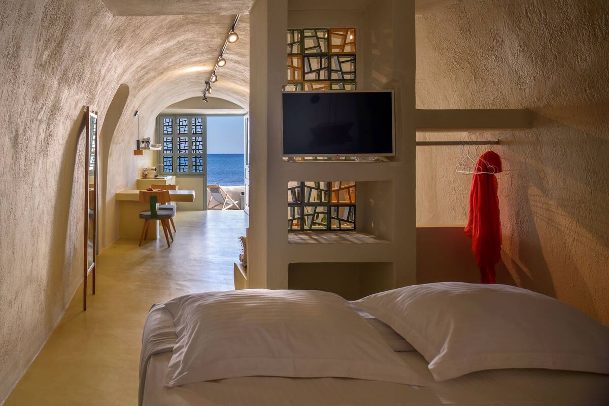 A bedroom with a large bed, a TV mounted on the wall, and a view of the sea through an arched doorway.