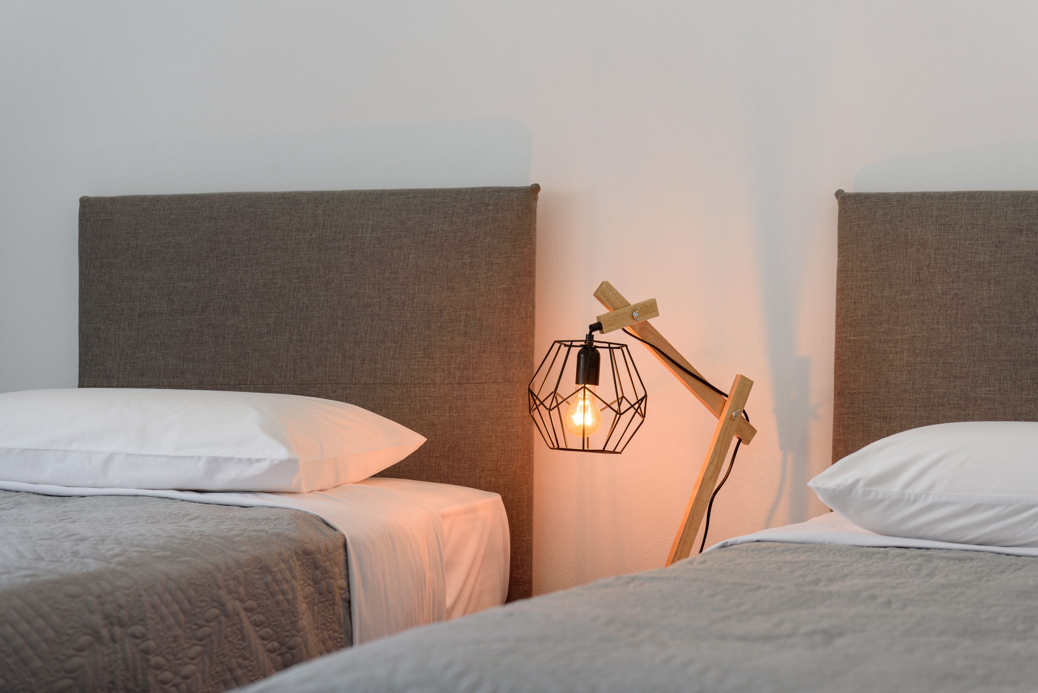 Et soveværelse med to senge, hver med sengegavl og hvide puder. En unik geometrisk lampe hænger på væggen mellem sengene.