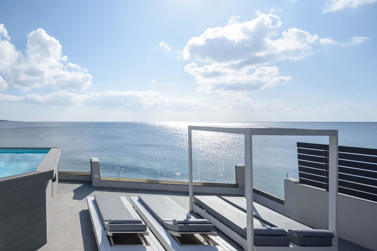 A rooftop pool area with a view of the sea, a lounge with sun loungers, and a staircase leading to the pool.