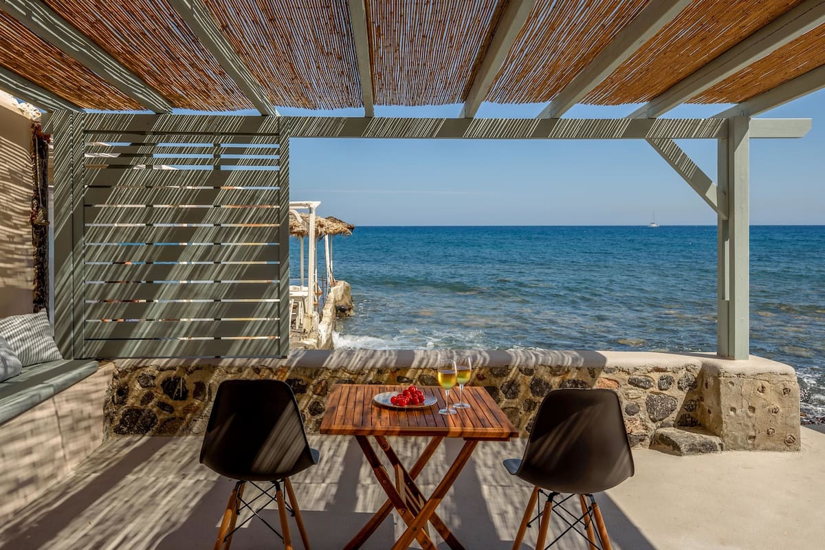 A terrace with a stone wall, a wooden table with two chairs, a glass of wine, and a plate of fruit.