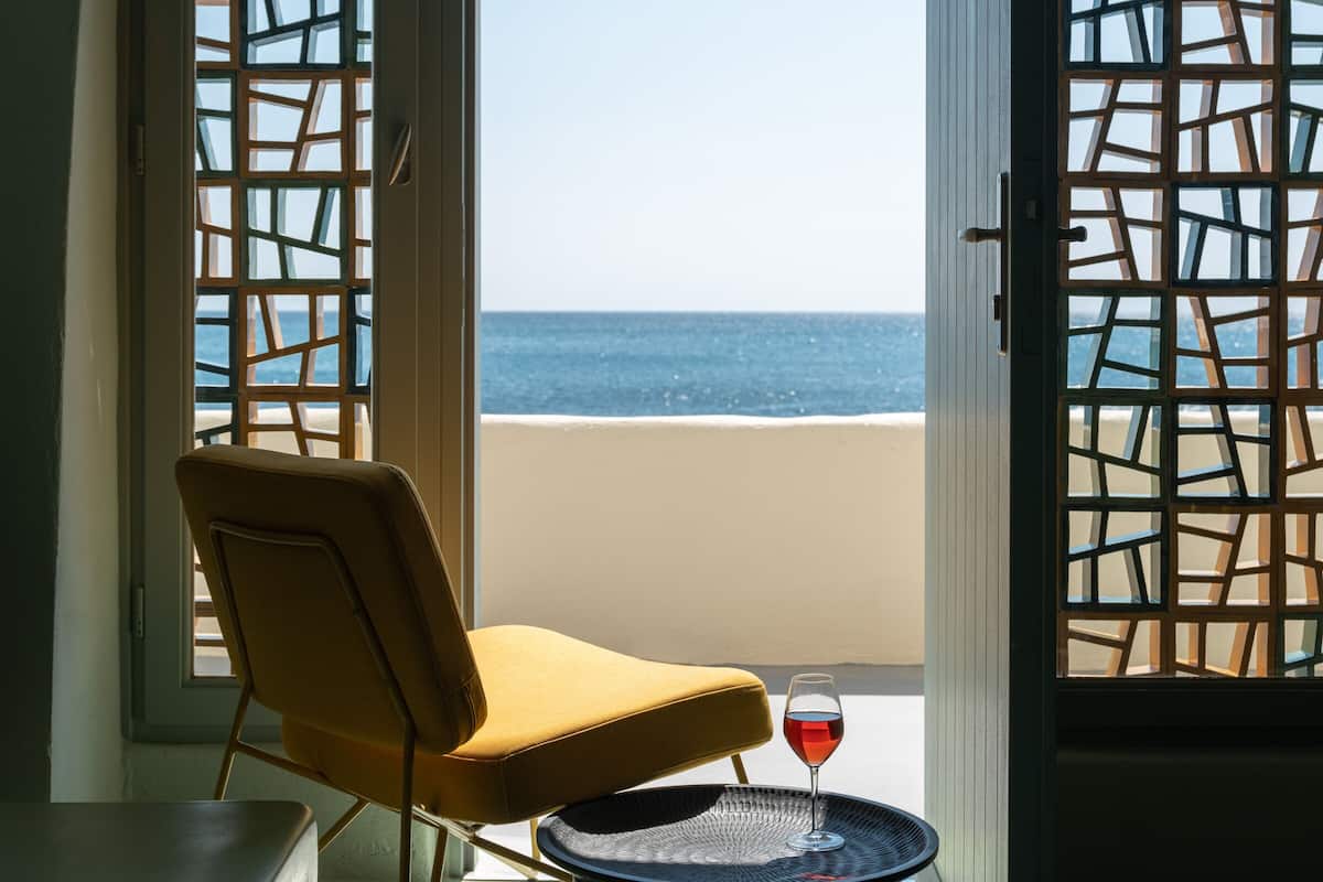 A room with a view of the beach, a chair, and a table with a glass of red wine.
