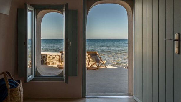 A room with a view of the sea through an open door, a beach chair, and a stone wall. - Sublime Villa & Caves (Santorini)