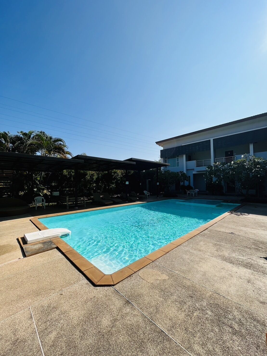 Una piscina rectangular con solárium y pavimento de baldosas, rodeada por un edificio con techo plano y una zona de patio cubierta.