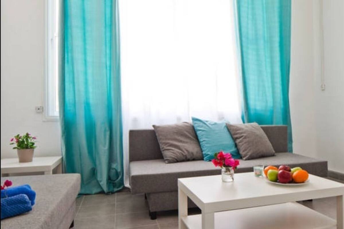 A modern living room with a grey sofa, teal curtains, a white coffee table with fruit, and a small plant on a side table.