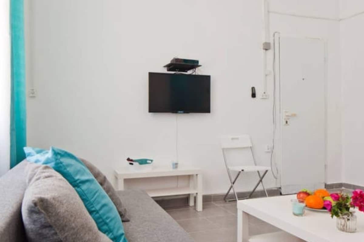 A modern living room with a flat-screen TV mounted on the wall, a white sofa with teal cushions, a small white table with a vase of flowers, and a white door in the background.