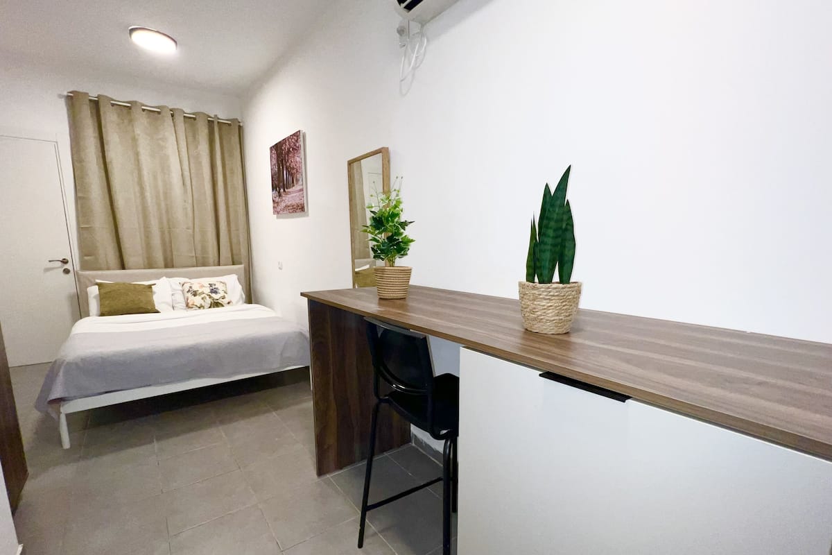 A small, clean room with a bed, a desk, and two potted plants.