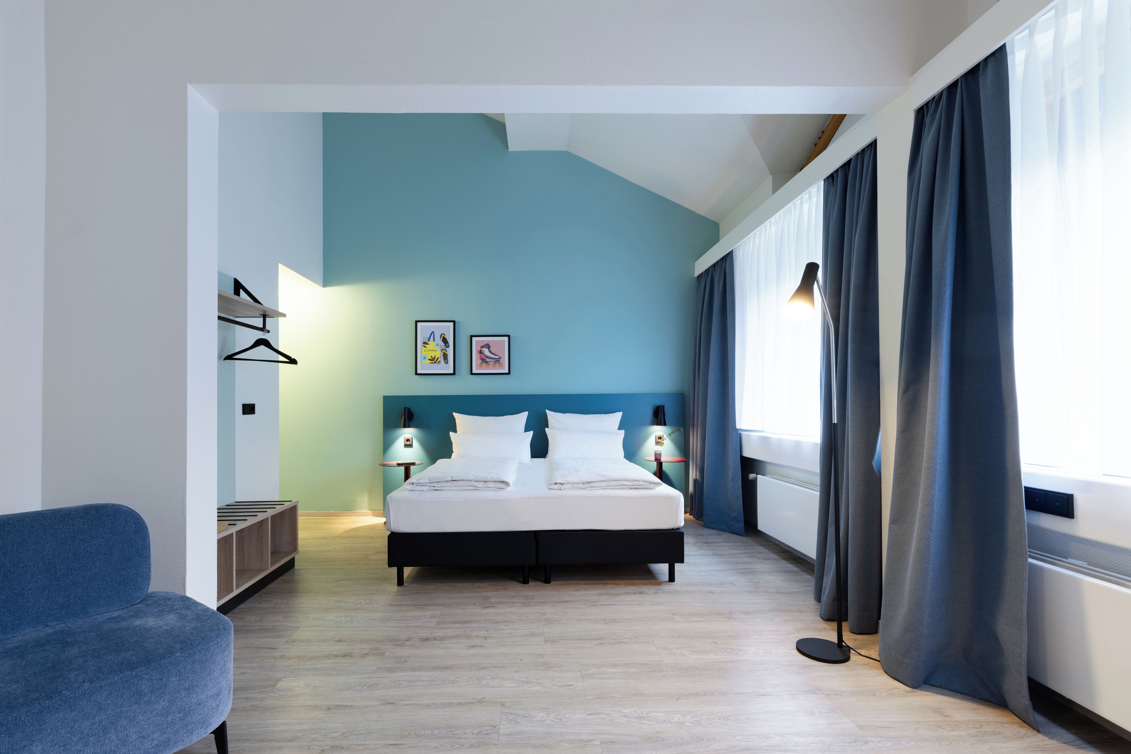 Een moderne hotelkamer met een bed, nachtlampjes, een blauwe muur en een raam met gordijnen.