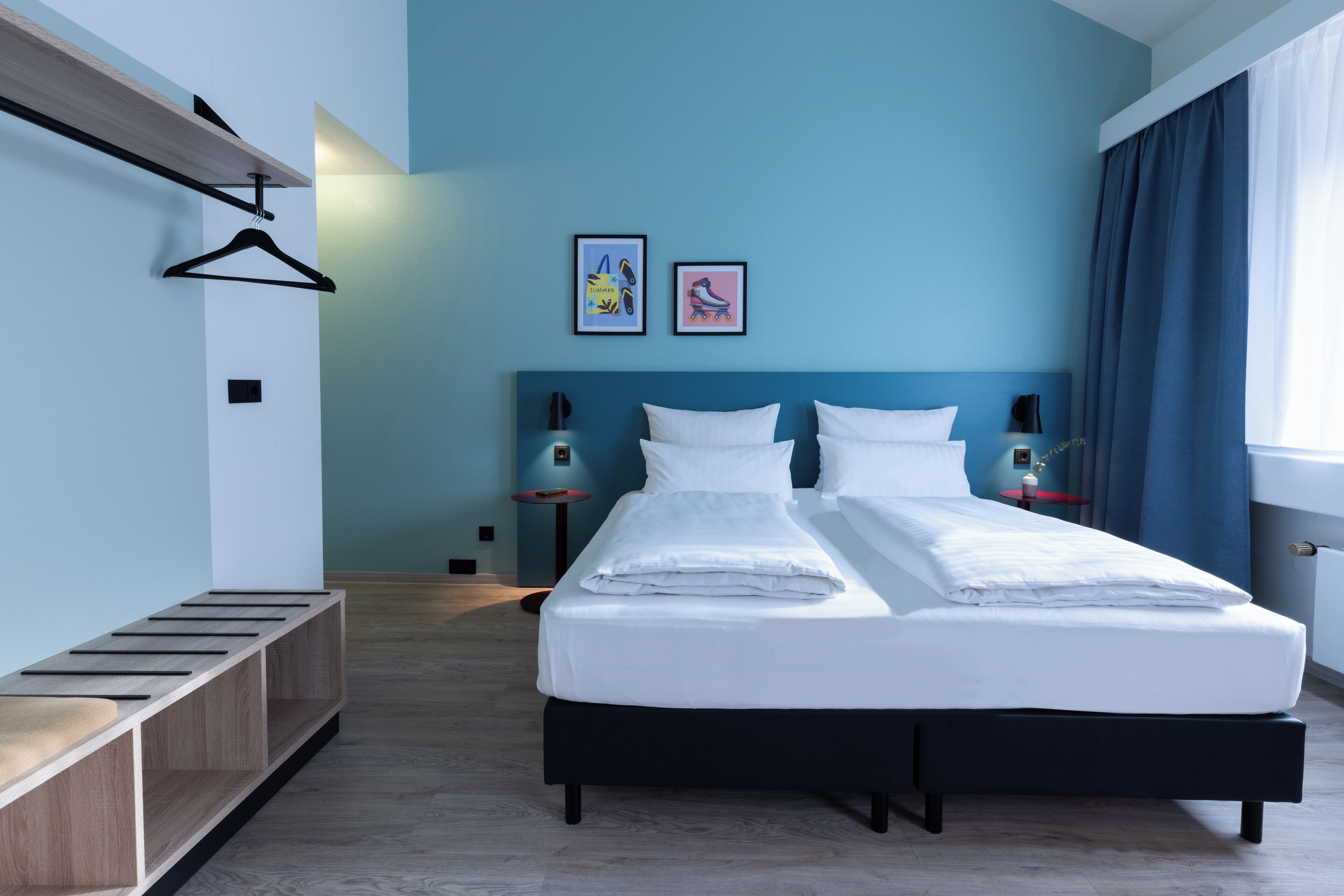 Una cama doble con sábanas blancas, un banco de madera, dos cuadros enmarcados y una mesita de noche con una lámpara.