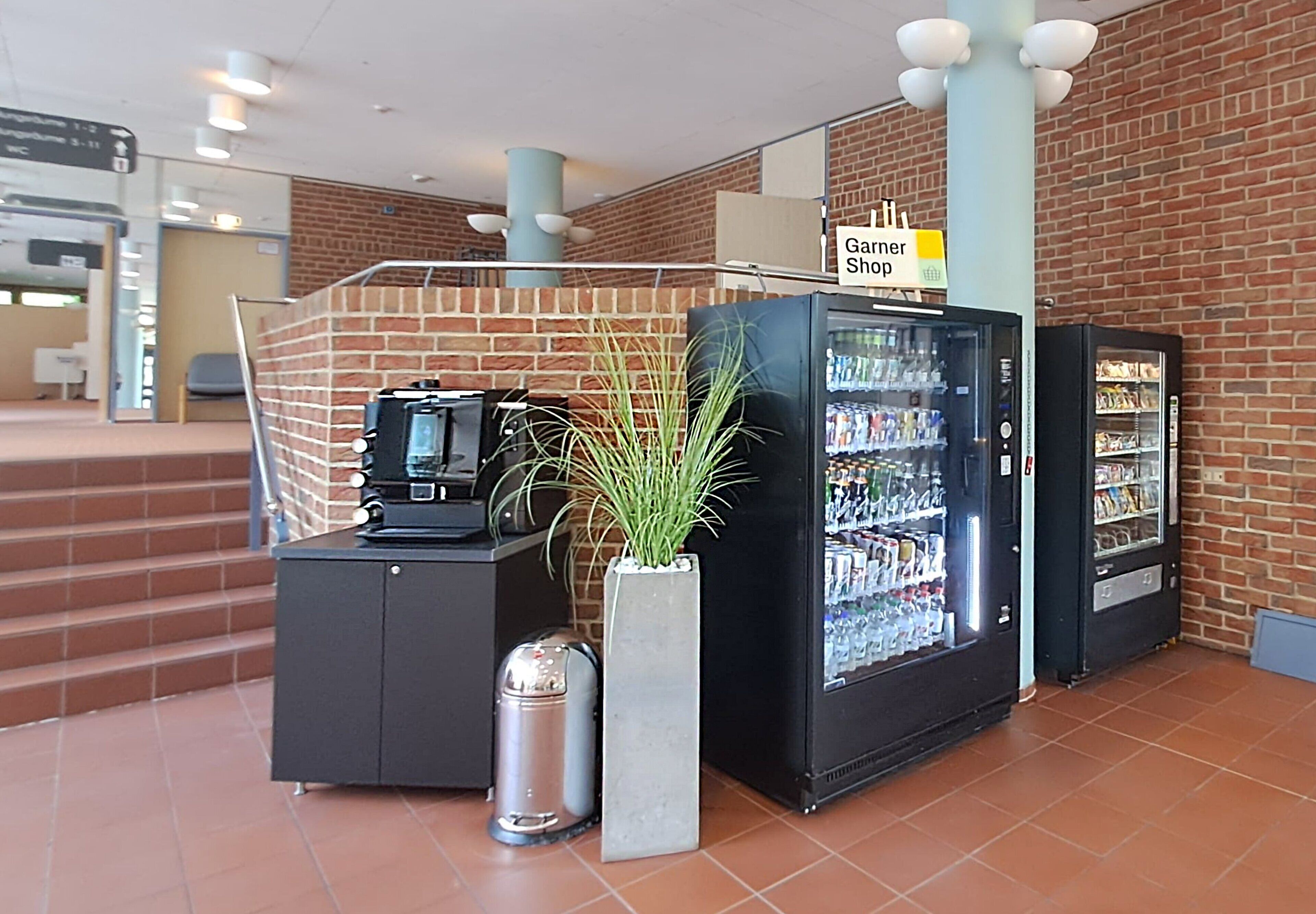 Un área de recepción con pared de ladrillo, un letrero de Garner Shop, máquinas expendedoras y un cubo de basura.