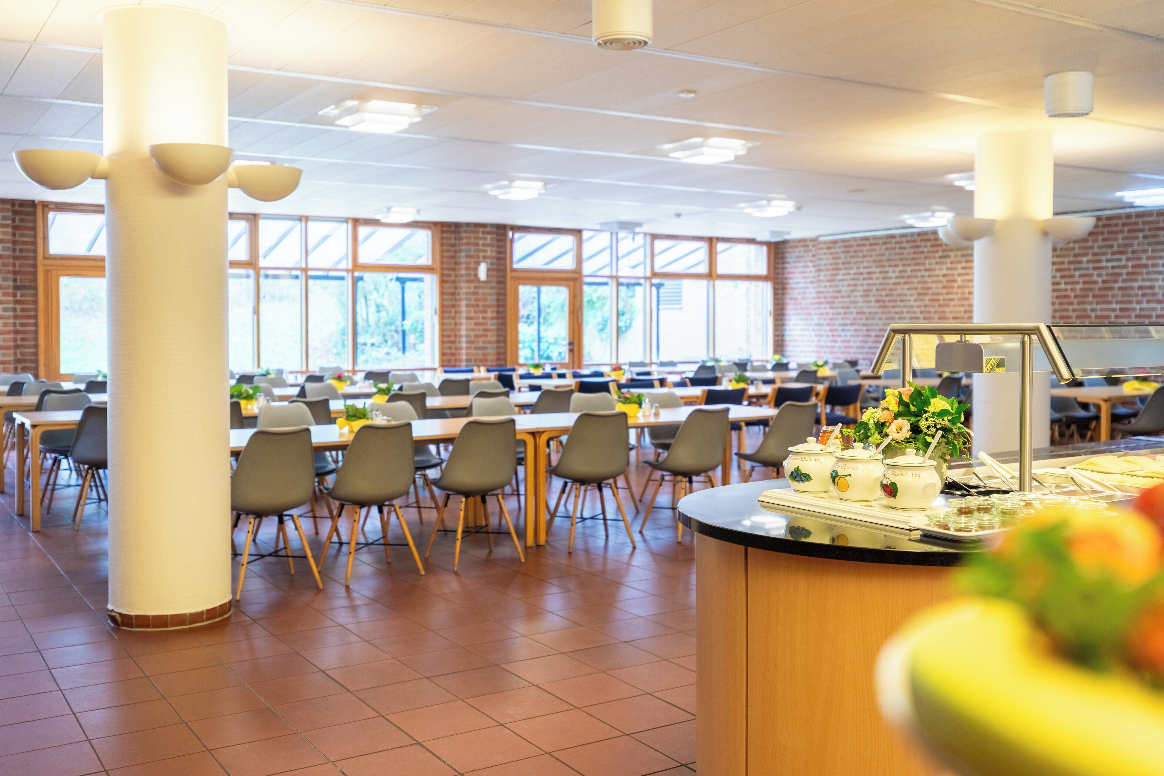 A spacious cafeteria with multiple tables and chairs, large windows, and a central serving area.