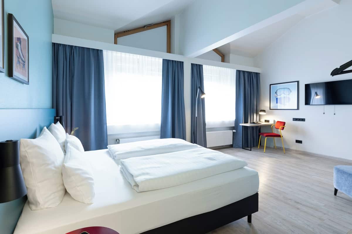 Een moderne hotelkamer met een groot bed, een bureau en een stoel. Er hangen gordijnen voor de ramen en een flatscreen televisie is aan de muur bevestigd.