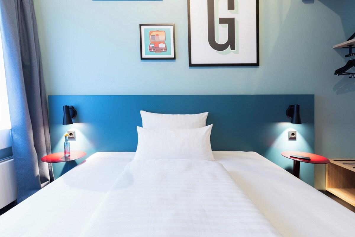 Un lit bien fait, avec du linge de lit blanc, deux tables de chevet avec des plateaux rouges et une tête de lit bleue.