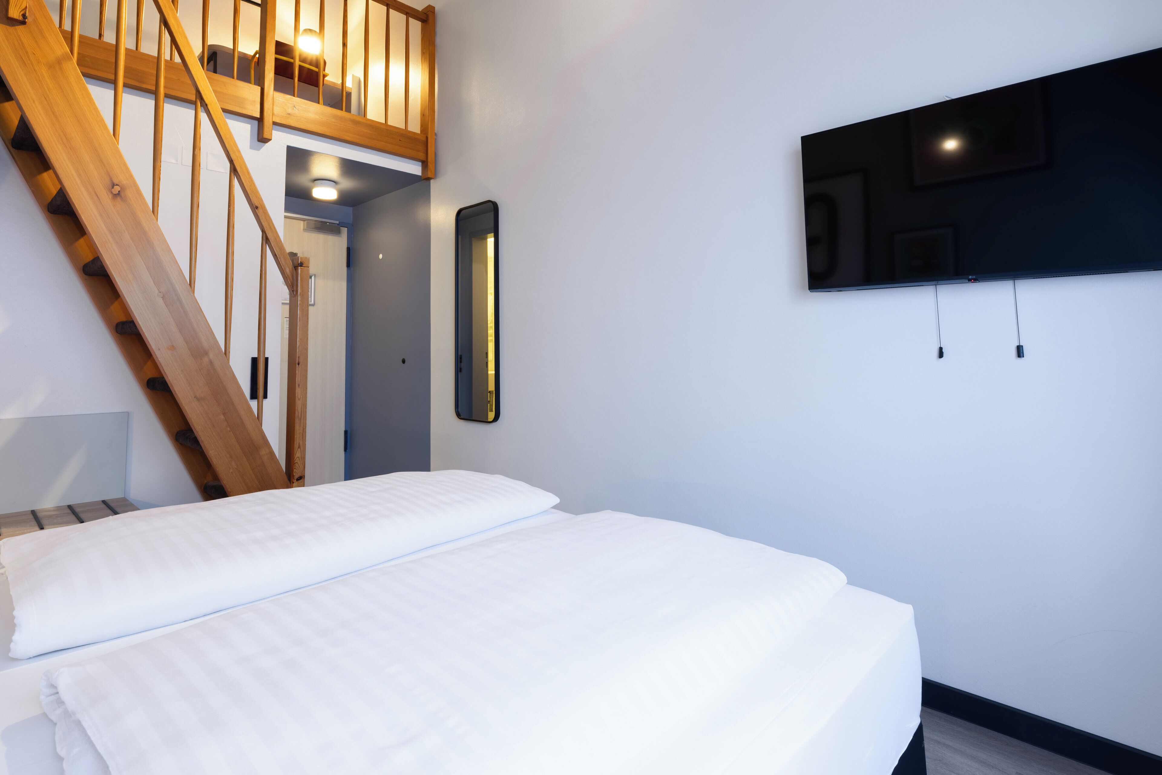 Un dormitorio con una escalera de madera, una cama con ropa de cama blanca, un televisor instalado en la pared y una mesita de noche.