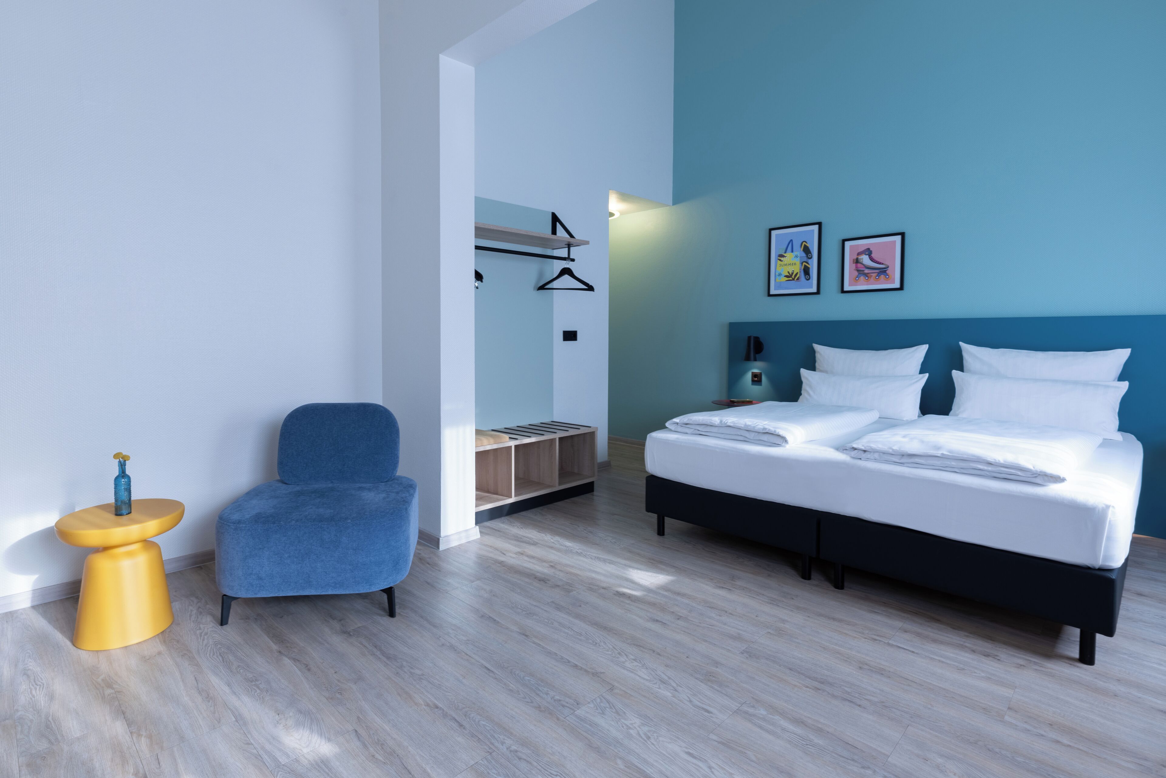 Une chambre à coucher moderne avec un lit, une chaise bleue, une petite table jaune et une étagère fixée au mur.