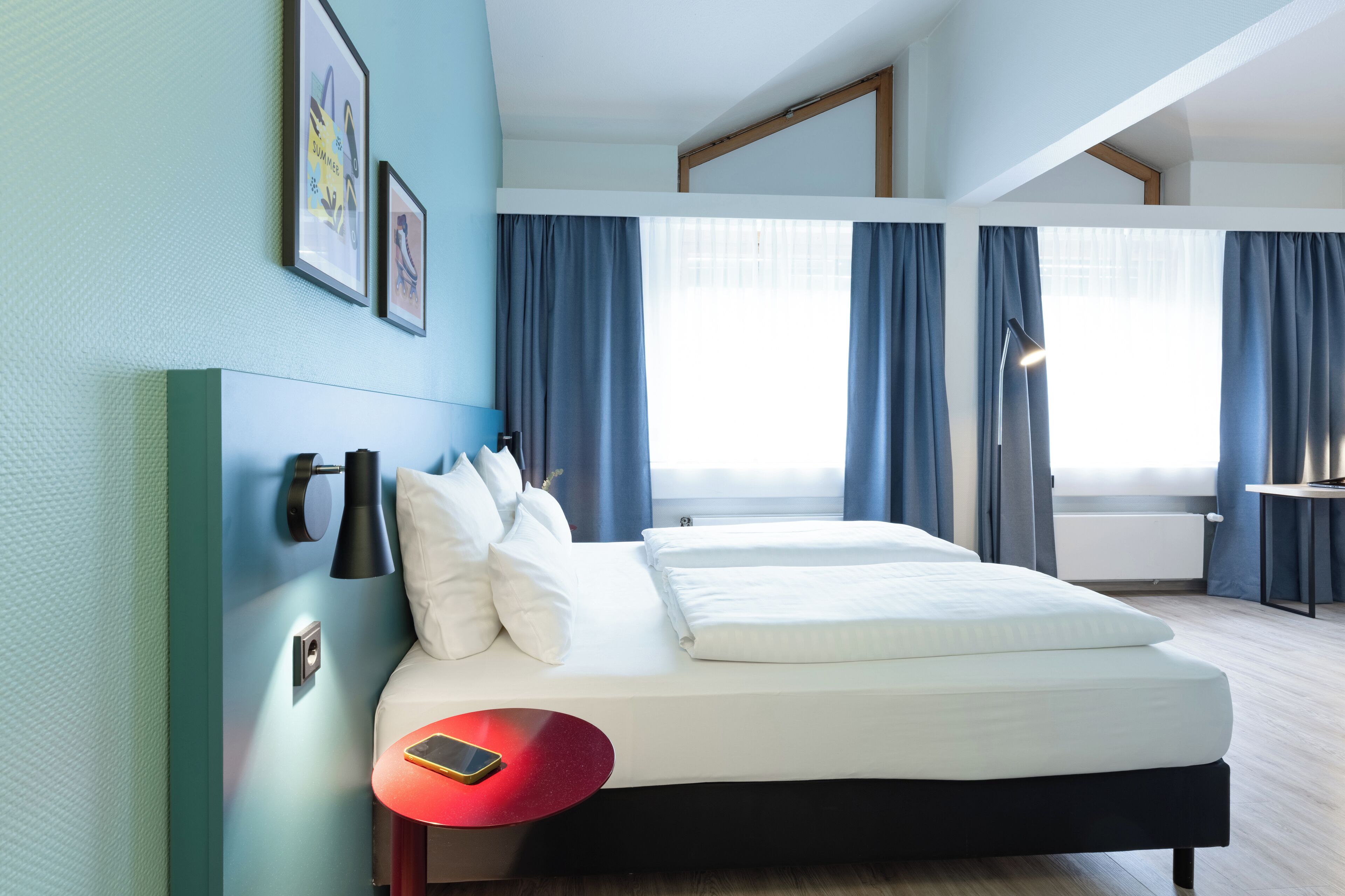 A modern hotel room with a bed, a red side table, a wall-mounted lamp, and a large window with curtains.