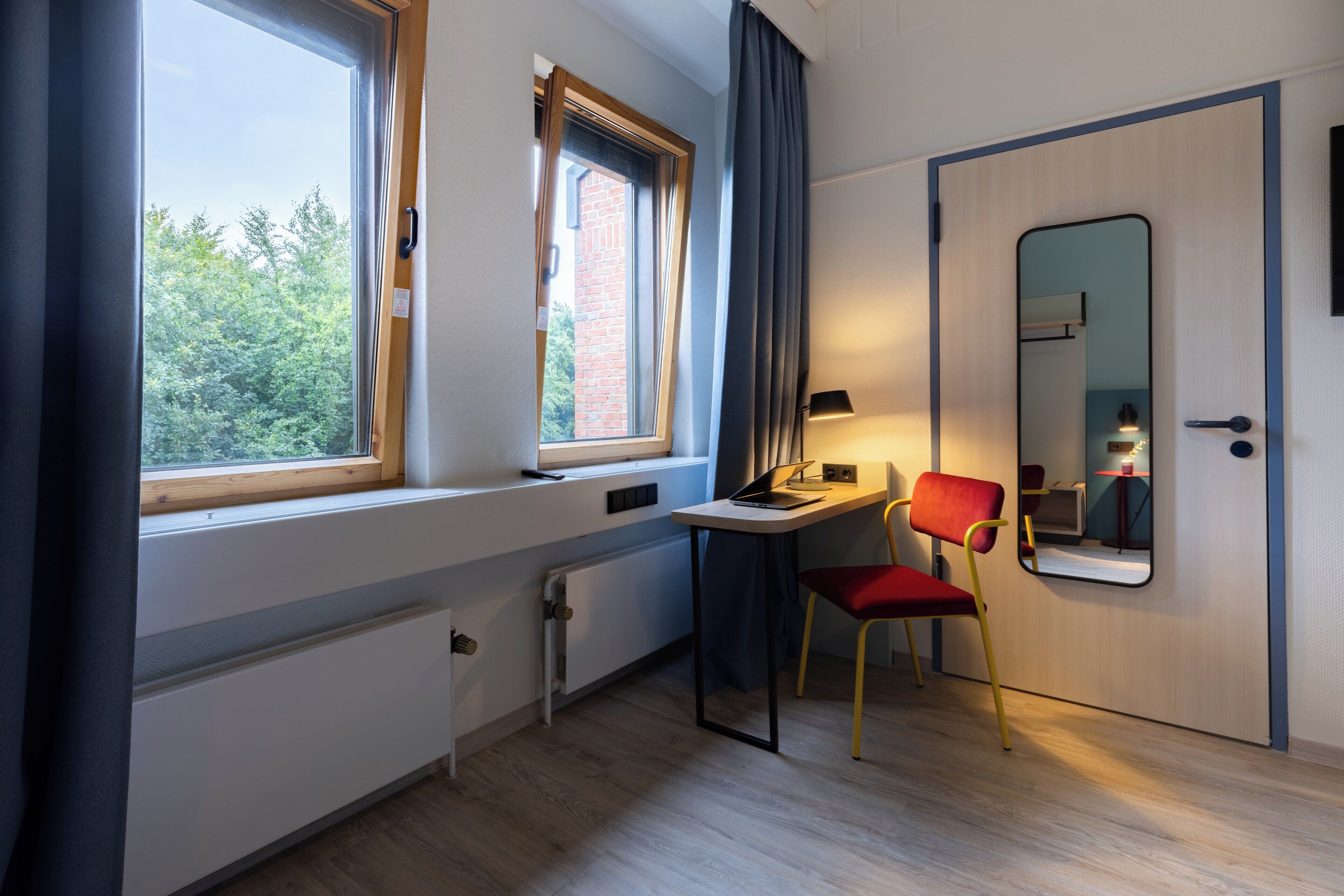 Een kamer met een bureau, stoel, raam, radiator en een deur met spiegel.