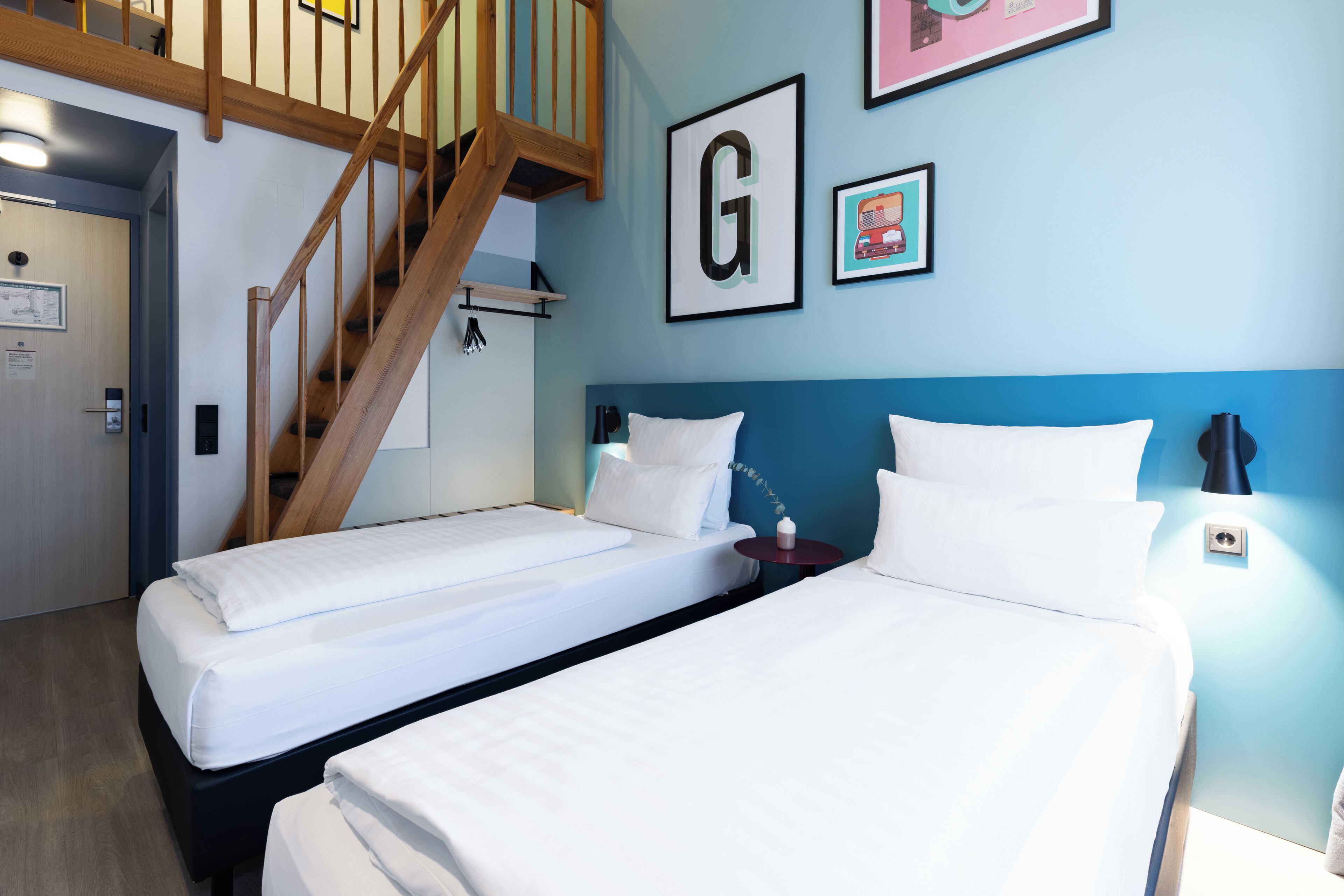 Ein Hotelzimmer mit zwei Einzelbetten, einer Holztreppe und einer blauen Wand mit einem gerahmten Buchstaben 'G'.