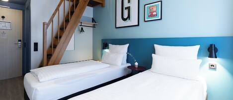 Ein Hotelzimmer mit zwei Einzelbetten, einer Holztreppe und einer blauen Wand mit einem gerahmten Buchstaben 'G'.