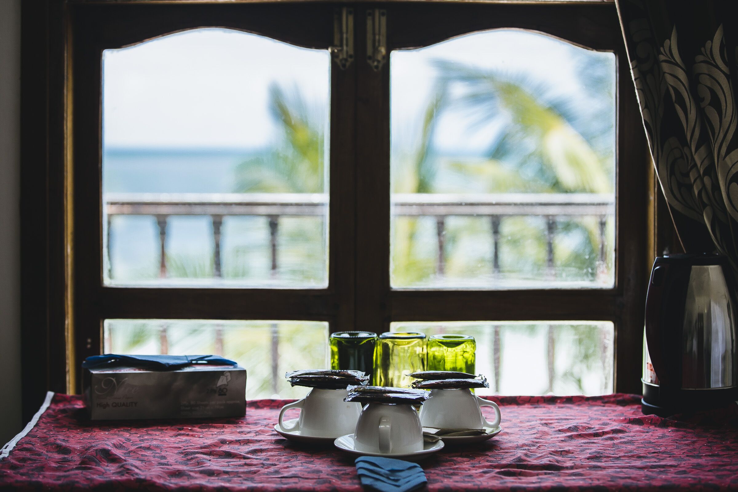 Una mesa con mantel rojo, una caja y cuatro vasos sobre platillos, con vista al mar a través de una ventana.