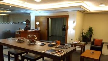 A hotel dining area with a long table, several chairs, a reception desk, and a potted plant.