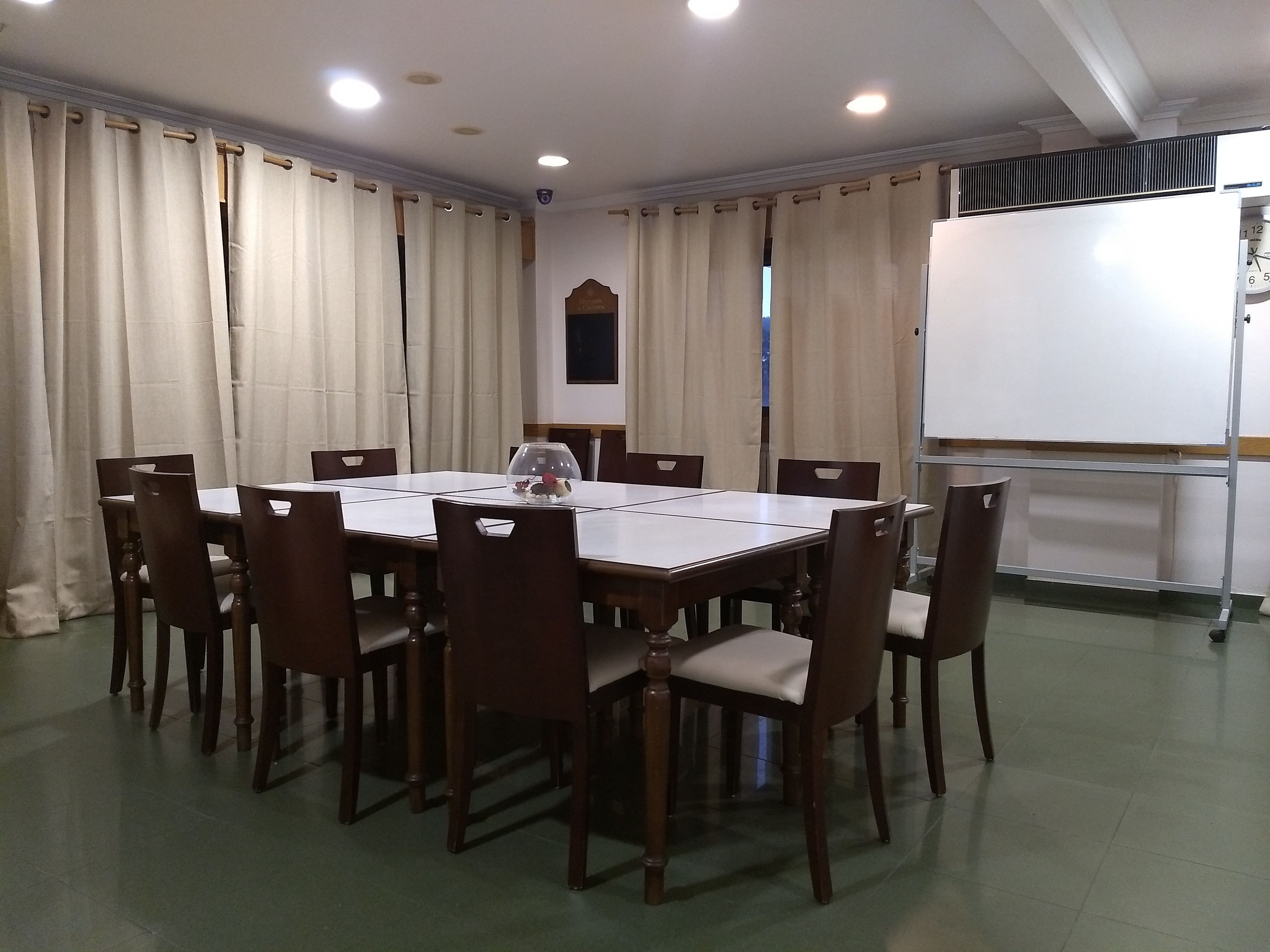 Une salle de conférence avec une table rectangulaire entourée de chaises en bois, un tableau blanc et des rideaux de couleur beige.