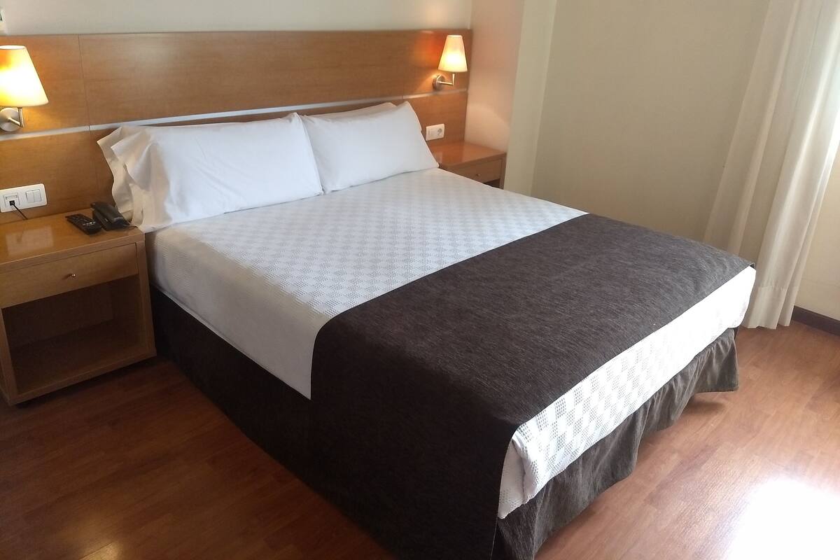 A neatly made bed with white and dark bedding, wooden headboard, bedside lamps, and a nightstand with a phone and remote.